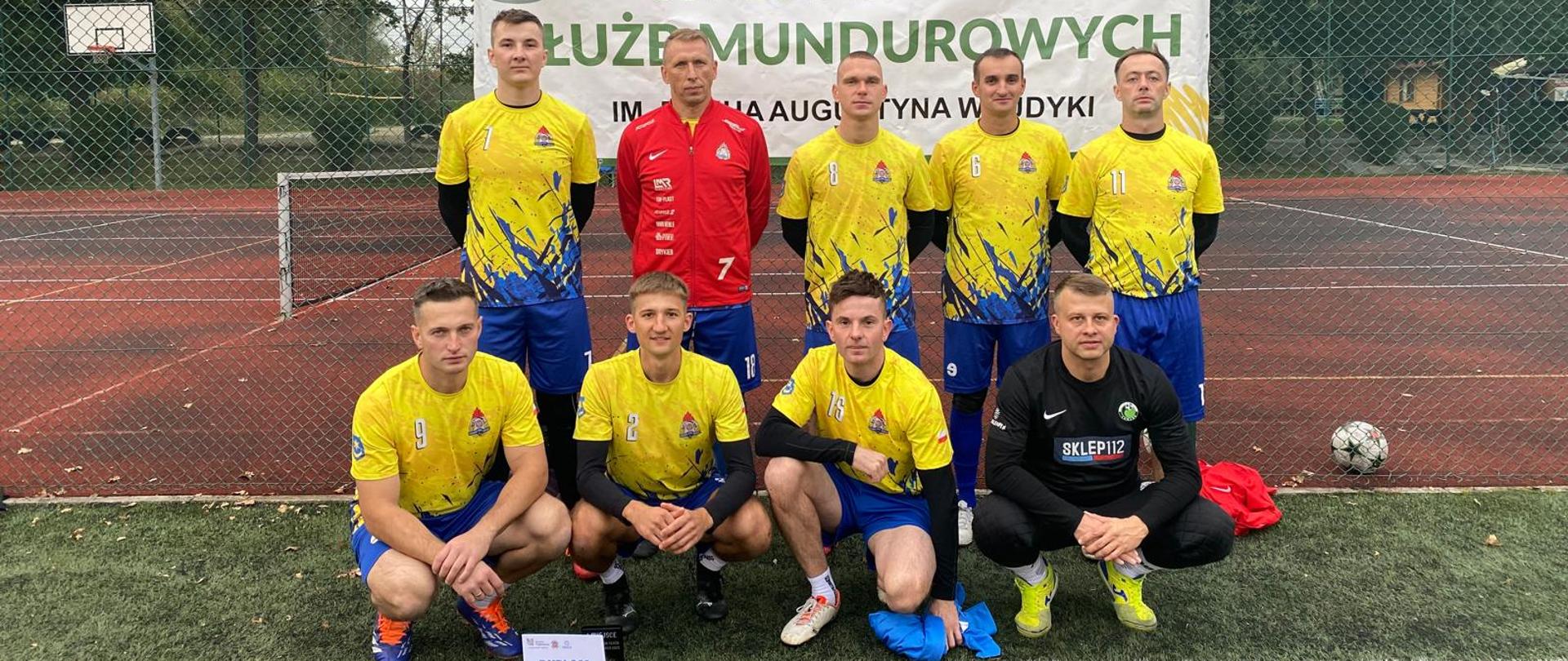 W dwóch rzędach stoi drużyna mężczyzn ubrani w większości w żółte podkoszulki i granatowe spodenki. Wszyscy ustawieni są na trawie, natomiast z tyłu widać boisko sportowe i drzewa