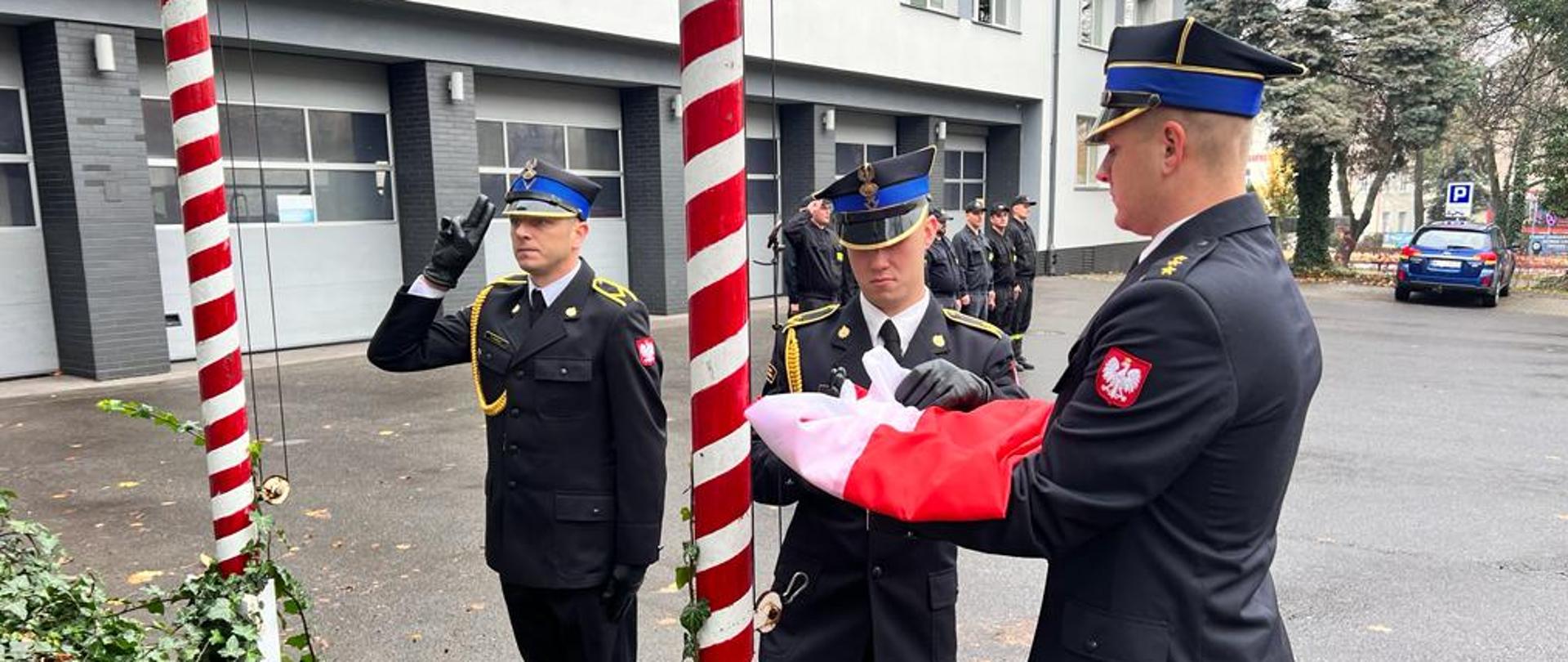 Zdjęcie przedstawia trzech funkcjonariuszy Państwowej Straży Pożarnej. Funkcjonariusze ubrani są w mundury galowe w kolorze ciemnym granatowym. Dwóch z nich przygotowuje flagę do wciągnięcia na maszt. Zdjęcie wykonane przed Jednostką Ratowniczo – Gaśniczą numer 1 w Radomiu.
