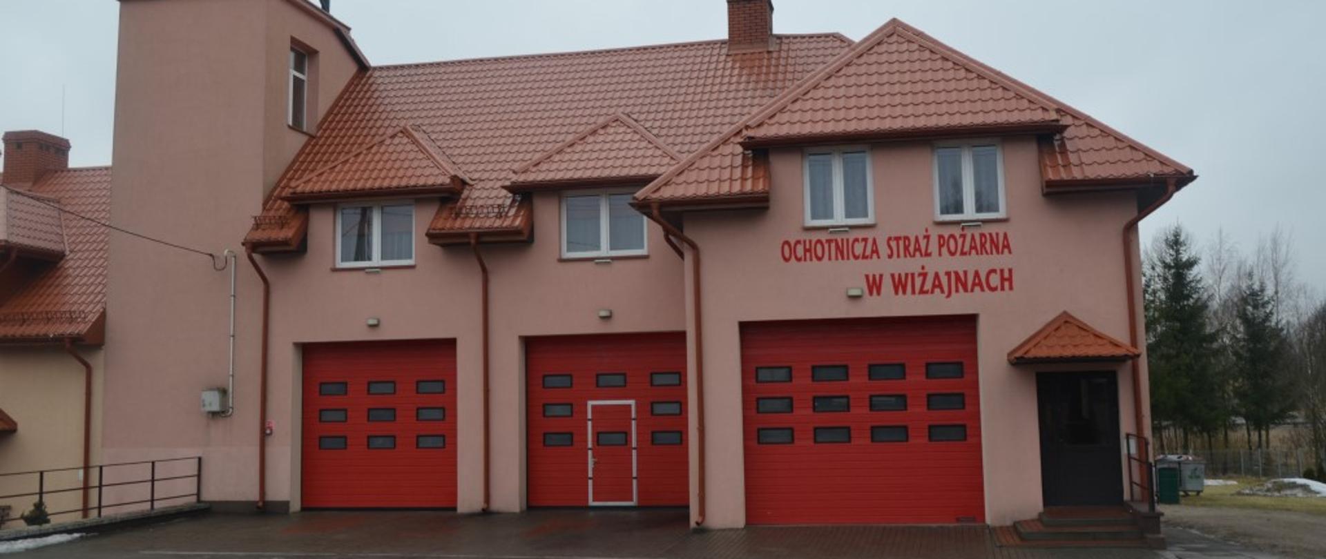 Widoczny budynek w kolorze elewacji jasno różowej z trzema bramami garażowymi koloru czerwonego. Z lewej strony wieża. Z prawej strony wejście do budynku. Dach koloru czerwonego. Podjazd wyłożony kostką. 