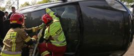 Na zdjęciu trzech strażaków demonstruje wykorzystanie podpór mechanicznych do szybkiej stabilizacji, używając ich do podparcia leżącego na boku samochodu osobowego. W tle grupa obserwująca