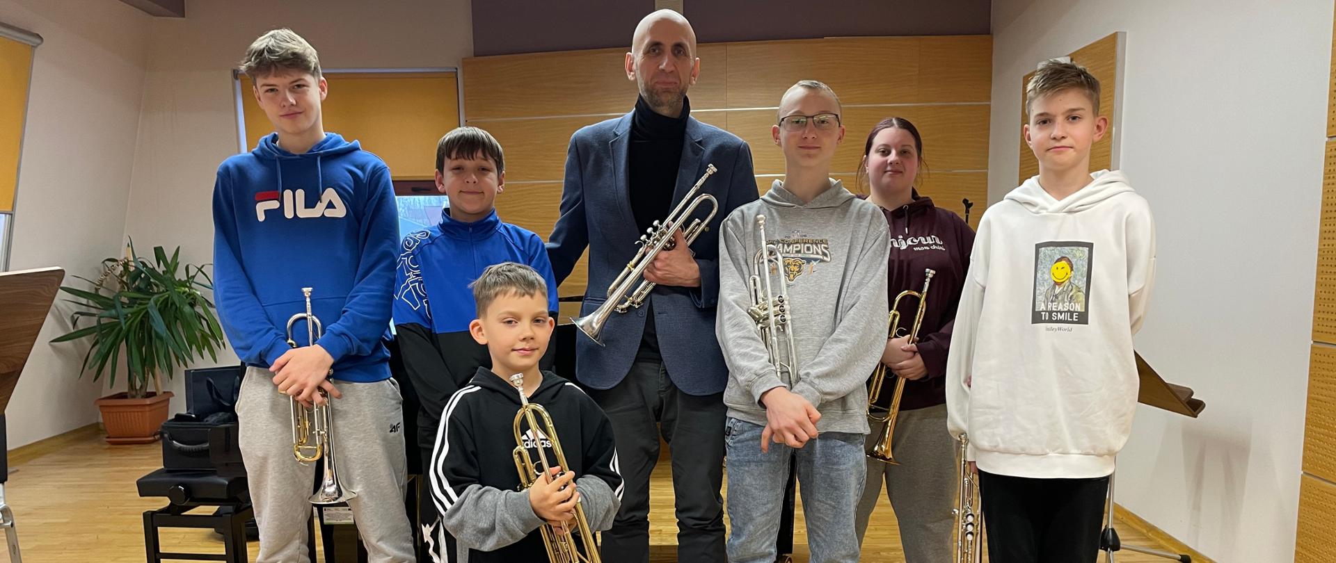 Zdjęcie grupowe. Uczniowie wraz z prowadzącym warsztaty stoją w zwartym szyku. Większość uczniów trzyma w swoich dłoniach instrumenty. Za nimi fortepian, pulpit, ława fortepianowa, na której futerał trąbkowy. W rogu sali za nimi palma doniczkowa, oraz dwa okna przysłonięte roletami.