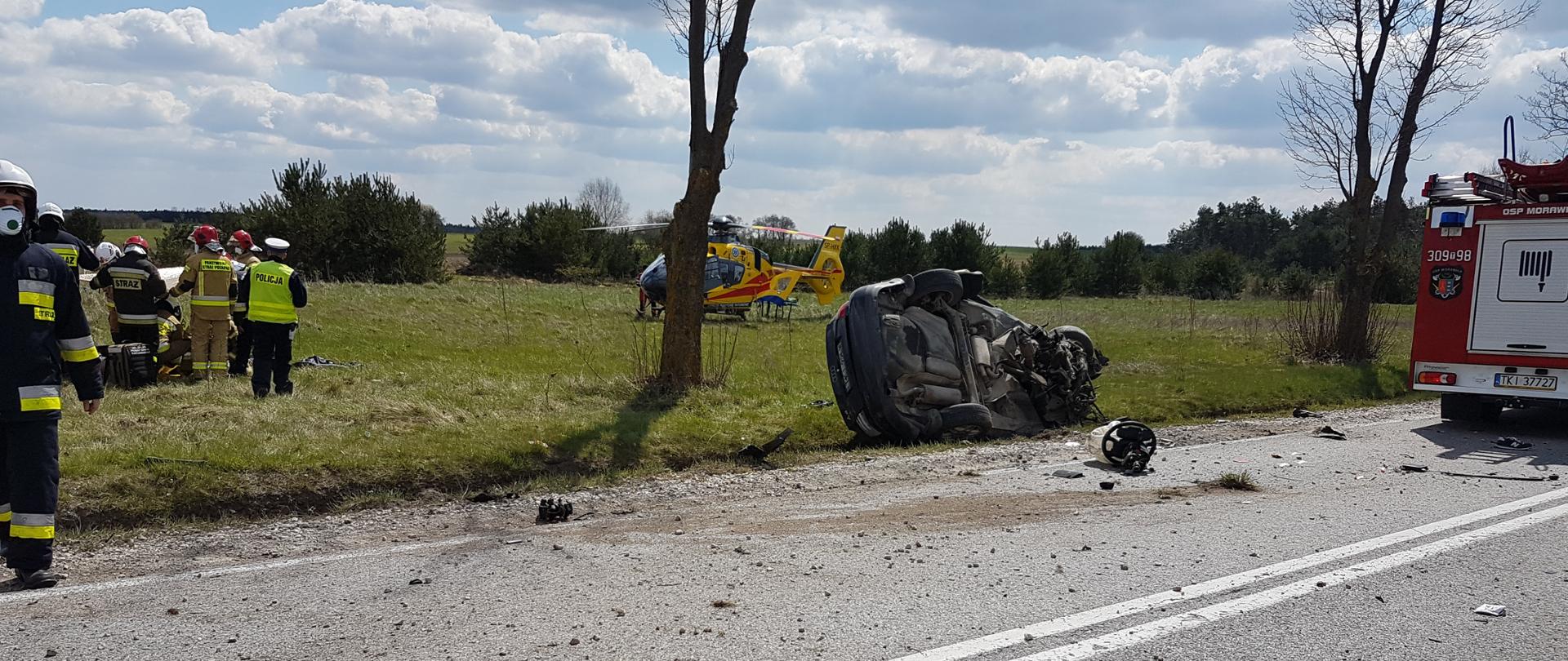 Zdjęcie przedstawia samochód osobowy, który znajduje się poza jezdnią drogi i jest przewrócony na dach. Wokół pojazdu widać porozrzucane liczne elementy karoserii. W oddali na łące znajduje się helikopter Lotniczego Pogotowia Ratunkowego, oraz służby ratownicze udzielające pomocy poszkodowanemu