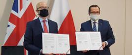 Na zdjęciu widać dwóch mężczyzn, którzy są ubrani w garnitury. Pokazują podpisane dokumenty. W tle, po lewej stronie widać flagę Wielkiej Brytanii, zaś po stronie prawej - flagę Polski. 