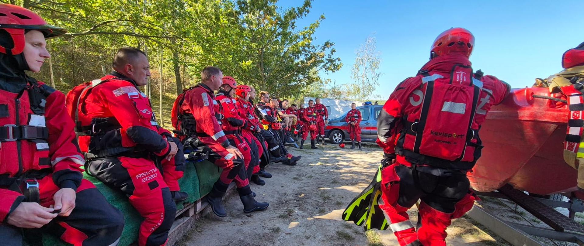 Strażacy ubrani w wodne skafandry wypornościowe siedzą na brzegu jeziora i wsłuchują się w prelekcję instruktora. Obok stoi łódź.