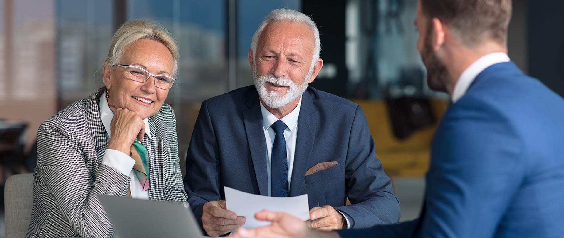 Na zdjęciu widoczna jest para w starszym wieku ubrana elegancko, która rozmawia z agentem ubezpieczeniowym ubranym w garnitur. U dołu zdjęcia widnieje logo ZUS