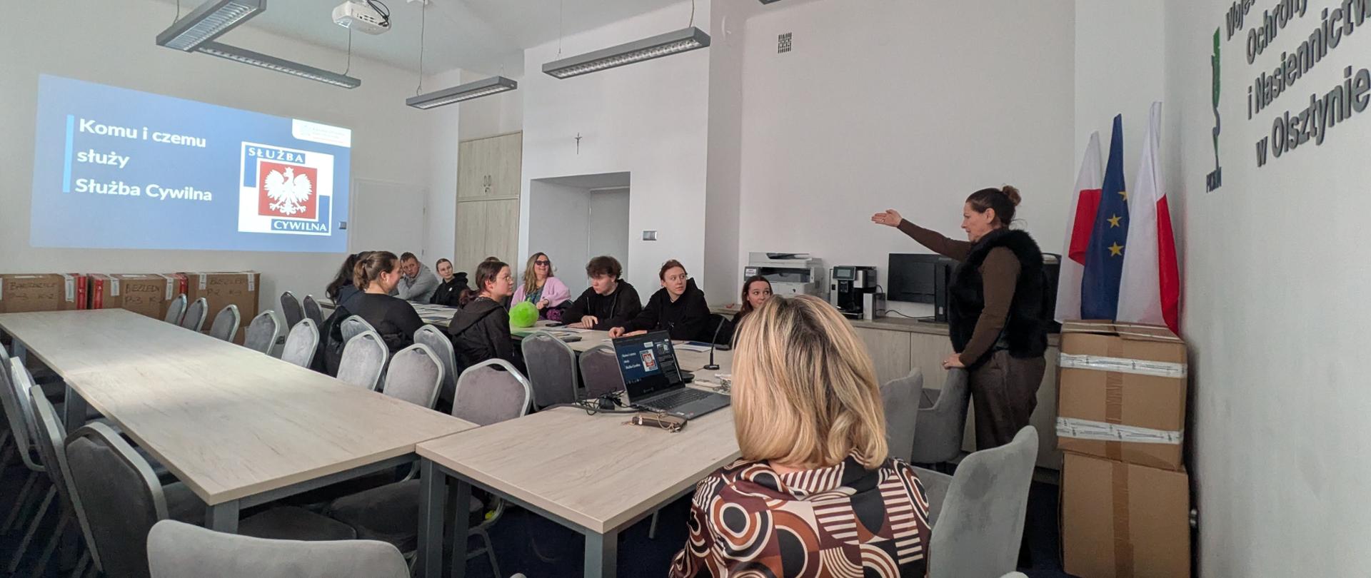 Grupa osób siedzi po obu stronach stołu i patrzy na stojącą po prawej stronie kobietę