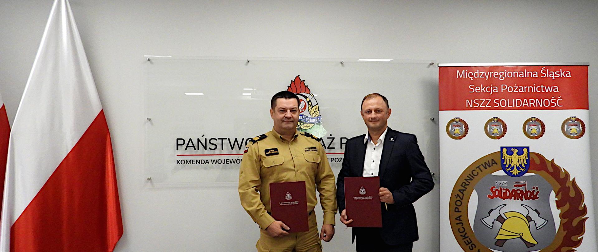 Na sali sztabowej w mundurze służbowym stoi śląski komendant wojewódzki PSP nadbryg. Jacek Kleszczewski oraz Przewodniczący Międzyregionalnej Śląskiej Sekcji Pożarnictwa NSZZ „Solidarność” Adam Rogoza w ciemnym garniturze. W rękach trzymają ozdobne, czerwone teczki z podpisanym porozumieniem. W tle na ścianie widoczne logo PSP. Po lewej stronie flagi państwowe. Po prawej baner z napisem Międzyregionalna Śląska Sekcja Pożarnictwa NSZZ „Solidarność” i logiem.