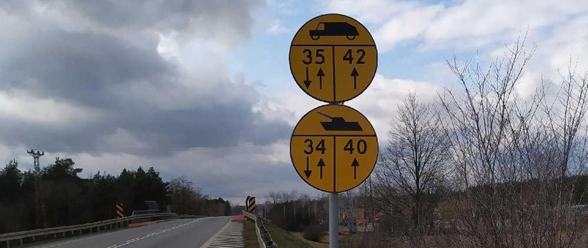 Znaki wojskowe przy drodze, po obu stronach drogi drzewa, w oddali zabudowania