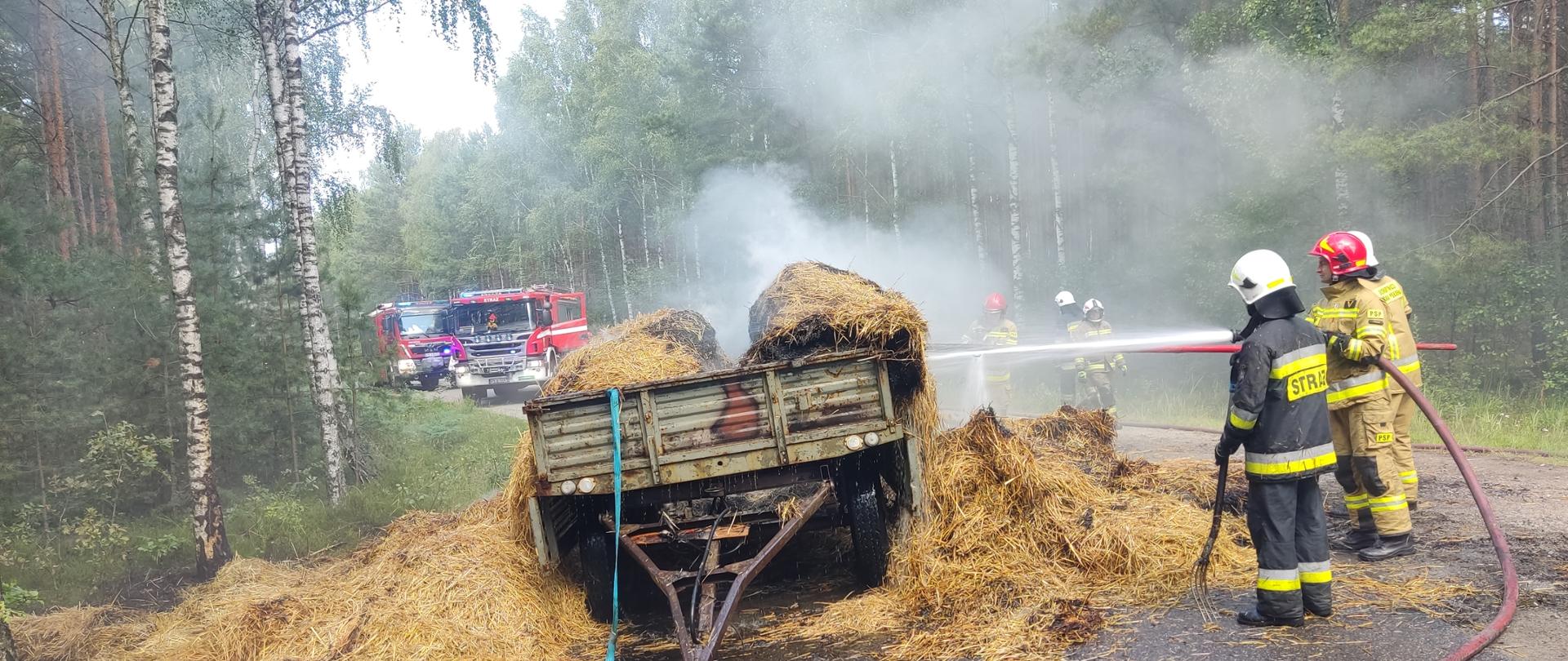 Na zdjęciu dymiąca się słoma na przyczepie rolniczej. Prądy wody podawane są przez umundurowanych ratowników. Zdjęcie w ciągu dnia. Na drodze.