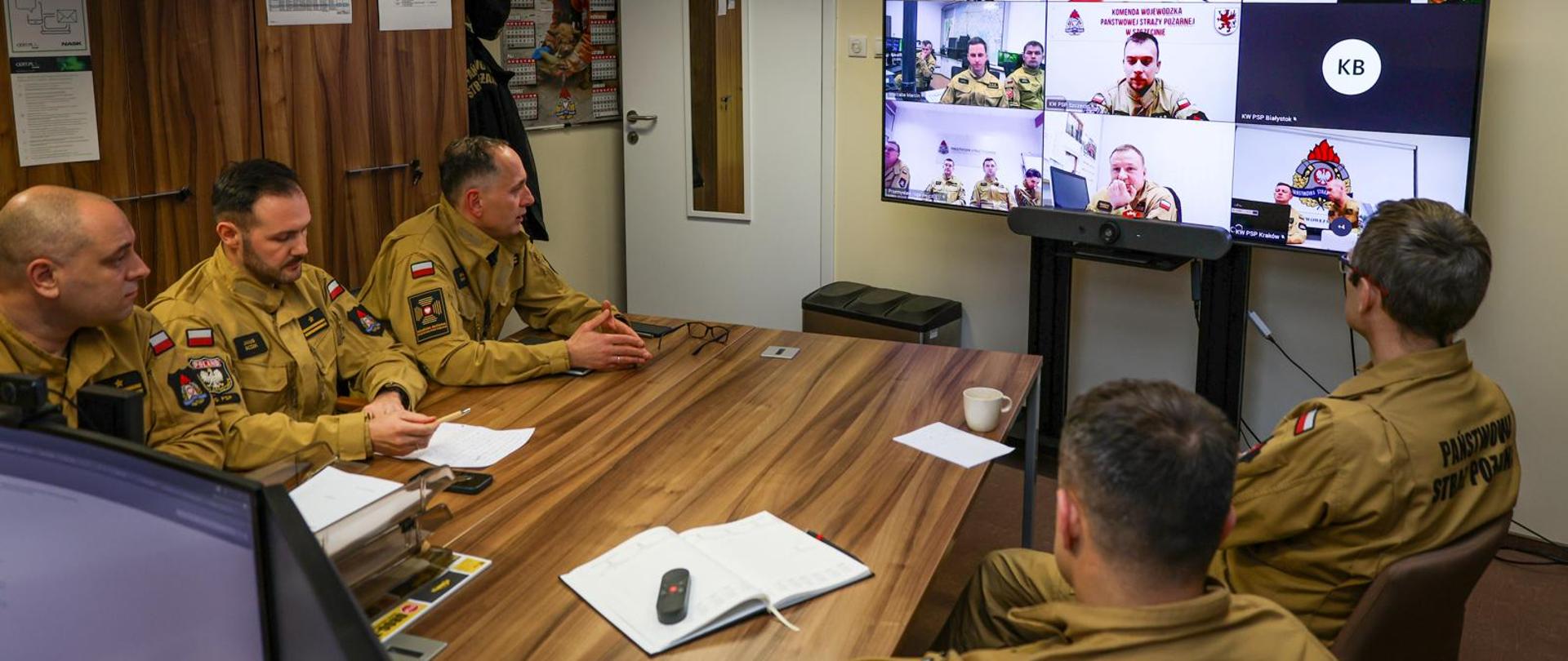 5 Funkcjonariuszy PSP siedzi przy stole w biurze, przed nimi monitor komputera, na którym widoczne są postacie innych strażaków - uczestników ćwiczeń sztabowych online