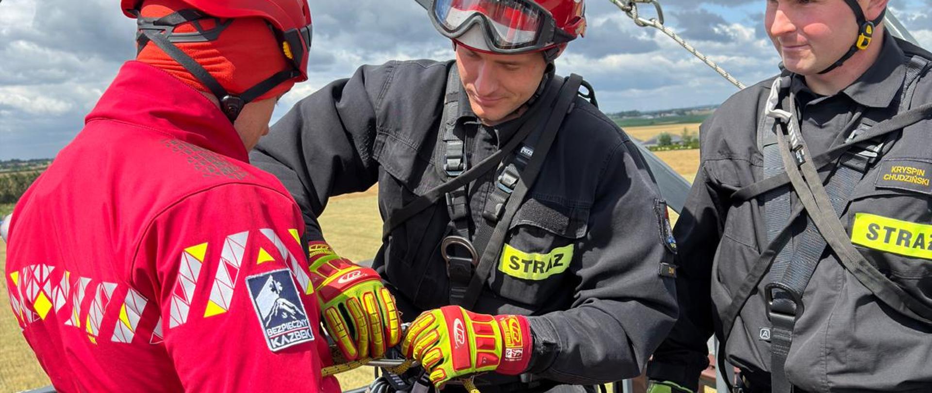 Trzech ratowników w kaskach ochronnych przygotowuje sprzęt linowy na wysokości podczas szkolenia na świeżym powietrzu. Strażak w czarnym ubraniu bojowym zapina karabinek przy uprzęży mężczyzny w czerwonej kurtce z logo „Bezpieczny Kazbek”, podczas gdy drugi strażak przygląda się ich działaniom.