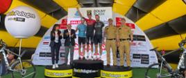 Trzech zawodników stoi na podium, jeden trzyma w ręku puchar uniesiony do góry, po prawo stoi dwóch strażaków w mundurach służbowych a po lewej stronie podium mężczyzna i kobieta w cywilnych ubraniach,