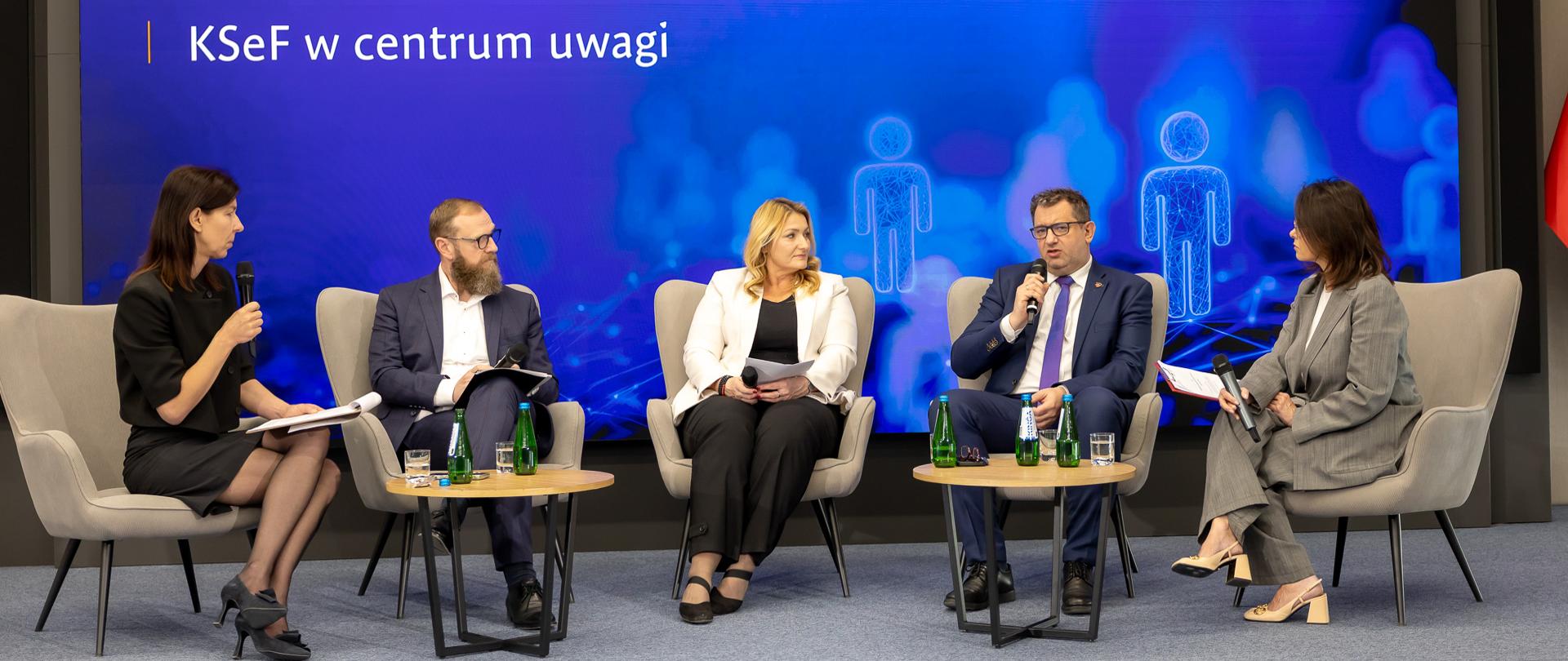 Marcin Łoboda Sekretarz Stanu w Ministerstwie Finansów przemawia do mikrofonu na tle ekranu informacyjnego wydarzenia.