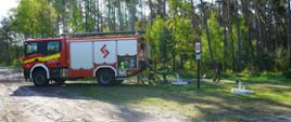 Na tle lasu stoi cięzki samochód pożarniczy. Za nim 2 strażaków, którzy sprawdzaja mozliwość poboru wody ze zbiornika podziemnego. Pomiędzy zbiornikami stoi tablica informacyjna - punkt czerpania wody. W poblizu zbiorników stoi lesniczy oraz pies.