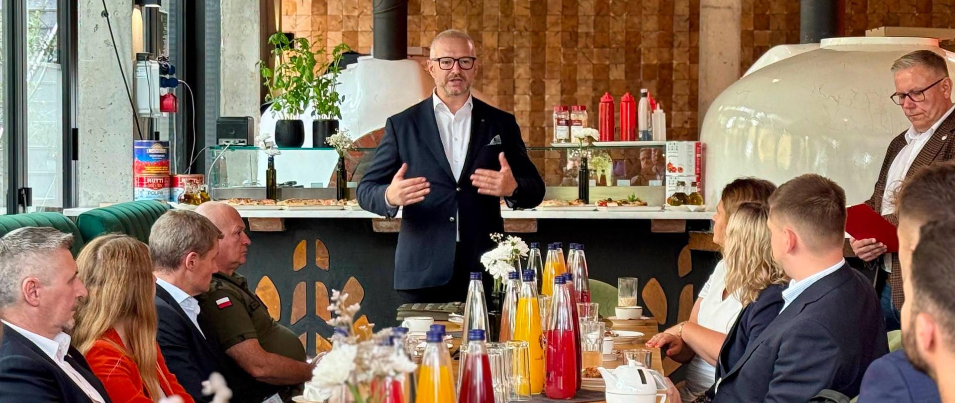 Wojewoda w garniturze stoi przy stole i przemawia do uczestników wydarzenia „Biznesowe Espresso”. Osoby siedzące przy stole z uwagą słuchają jego wystąpienia.