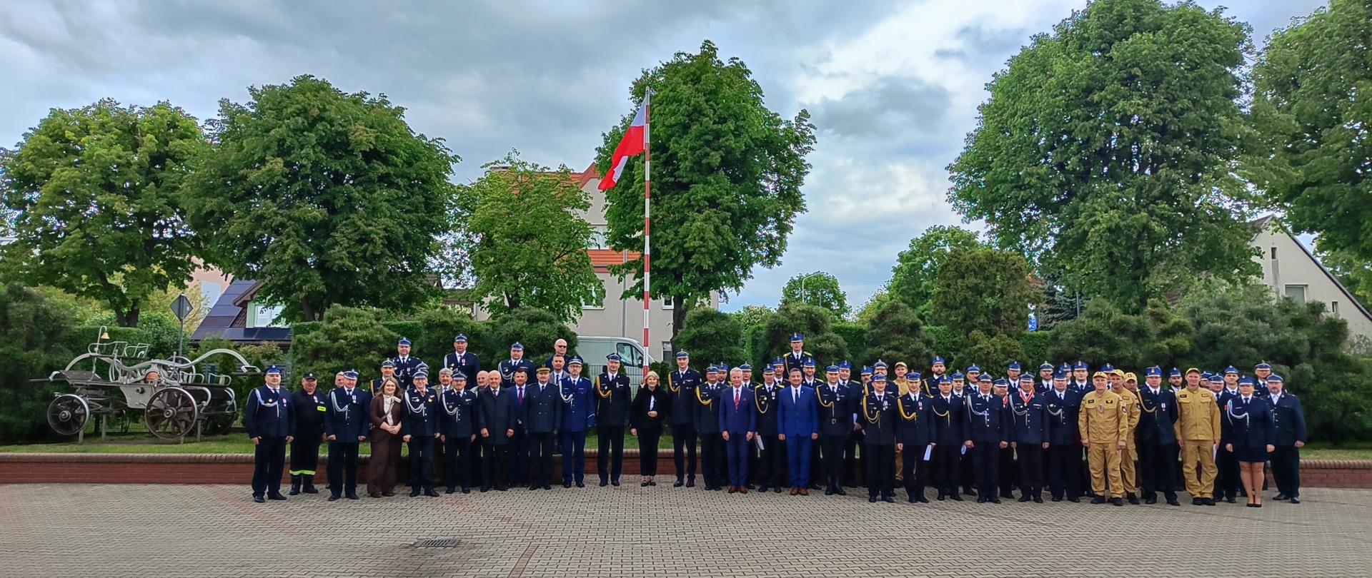 Grupowe zdjęcie kilkudziesięciu strażaków w mundurach galowych oraz osób cywilnych, pozujących na wybrukowanym placu. W centralnej części tła widoczny jest wysoki maszt z polską flagą, a po lewej stronie znajduje się zabytkowa, konna sikawka strażacka. Całość kompozycji dopełniają gęste, zielone drzewa pod lekko zachmurzonym niebem.