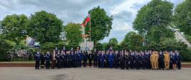Grupowe zdjęcie kilkudziesięciu strażaków w mundurach galowych oraz osób cywilnych, pozujących na wybrukowanym placu. W centralnej części tła widoczny jest wysoki maszt z polską flagą, a po lewej stronie znajduje się zabytkowa, konna sikawka strażacka. Całość kompozycji dopełniają gęste, zielone drzewa pod lekko zachmurzonym niebem.