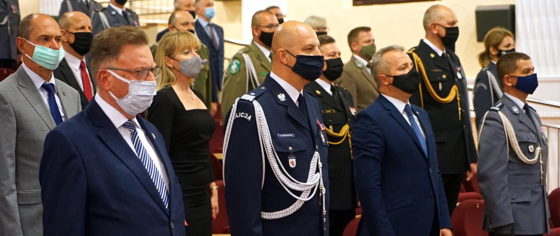 Na zdjęciu widać stojących w auli policjantów i zaproszonych gości. Policjanci mają na sobie umundurowanie wyjściowe. W pierwszej ławie obok komendanta wojewódzkiego policji stoi wojewoda kujawsko-pomorski oraz wicemarszałek województwa kujawsko-pomorskiego. W drugim rzędzie widać zastępcę kujawsko-pomorskiego komendanta wojewódzkiego Państwowej Straży Pożarnej oraz innych zaproszonych gości.