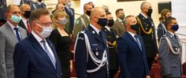 Na zdjęciu widać stojących w auli policjantów i zaproszonych gości. Policjanci mają na sobie umundurowanie wyjściowe. W pierwszej ławie obok komendanta wojewódzkiego policji stoi wojewoda kujawsko-pomorski oraz wicemarszałek województwa kujawsko-pomorskiego. W drugim rzędzie widać zastępcę kujawsko-pomorskiego komendanta wojewódzkiego Państwowej Straży Pożarnej oraz innych zaproszonych gości.