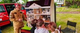 Strażak opowiada dzieciom o zagrożeniach. W tle mobilny symulator zagrożeń. Dalej z lewej strony stoi samochód straży pożarnej.