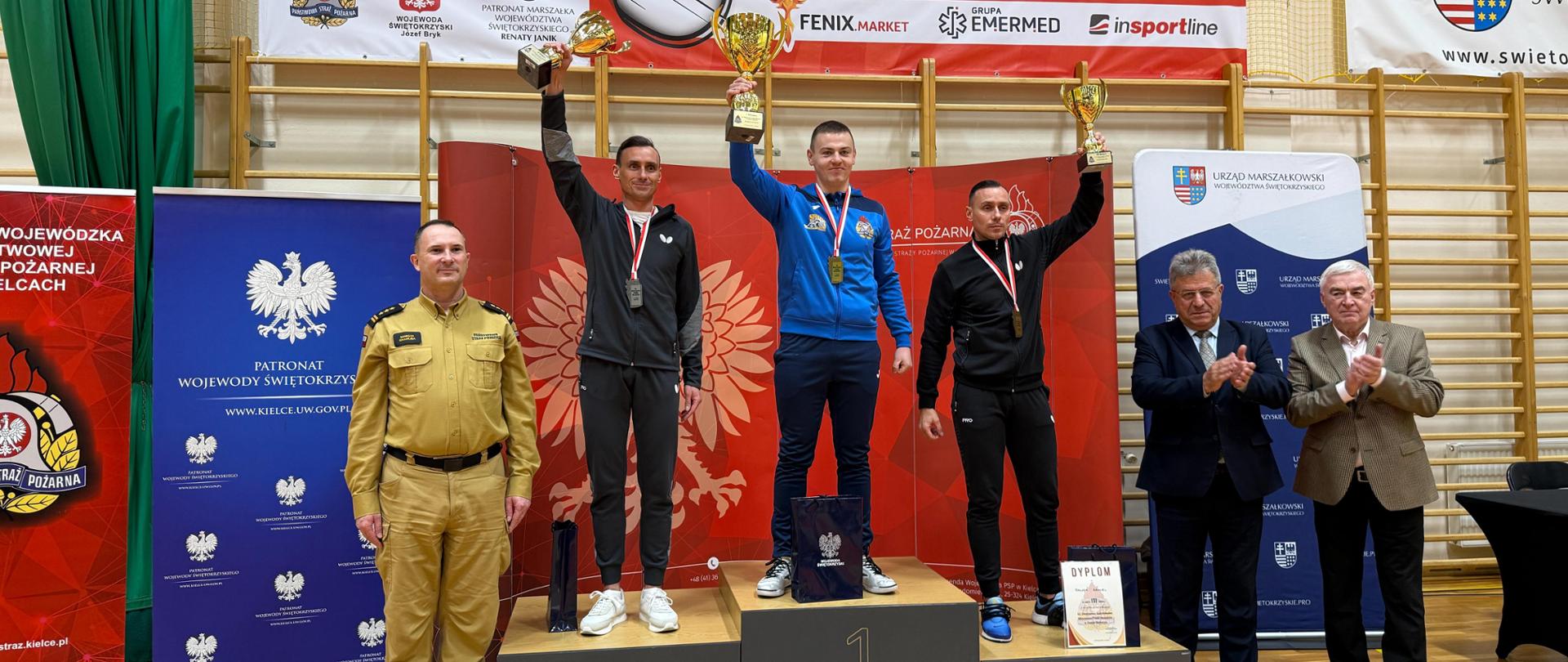Zwycięzcy rywalizacji indywidualnej stoją na podium podczas Mistrzostw Polski Strażaków PSP w Tenisie Stołowym. Zawodnik na pierwszym miejscu trzyma złoty puchar, podobnie jak zdobywcy drugiego i trzeciego miejsca. Z lewej strony stoi funkcjonariusz PSP w mundurze wyjściowym, a z prawej przedstawiciele władz wojewódzkich biją brawo. W tle widoczne są banery organizatorów oraz duży banner wydarzenia.