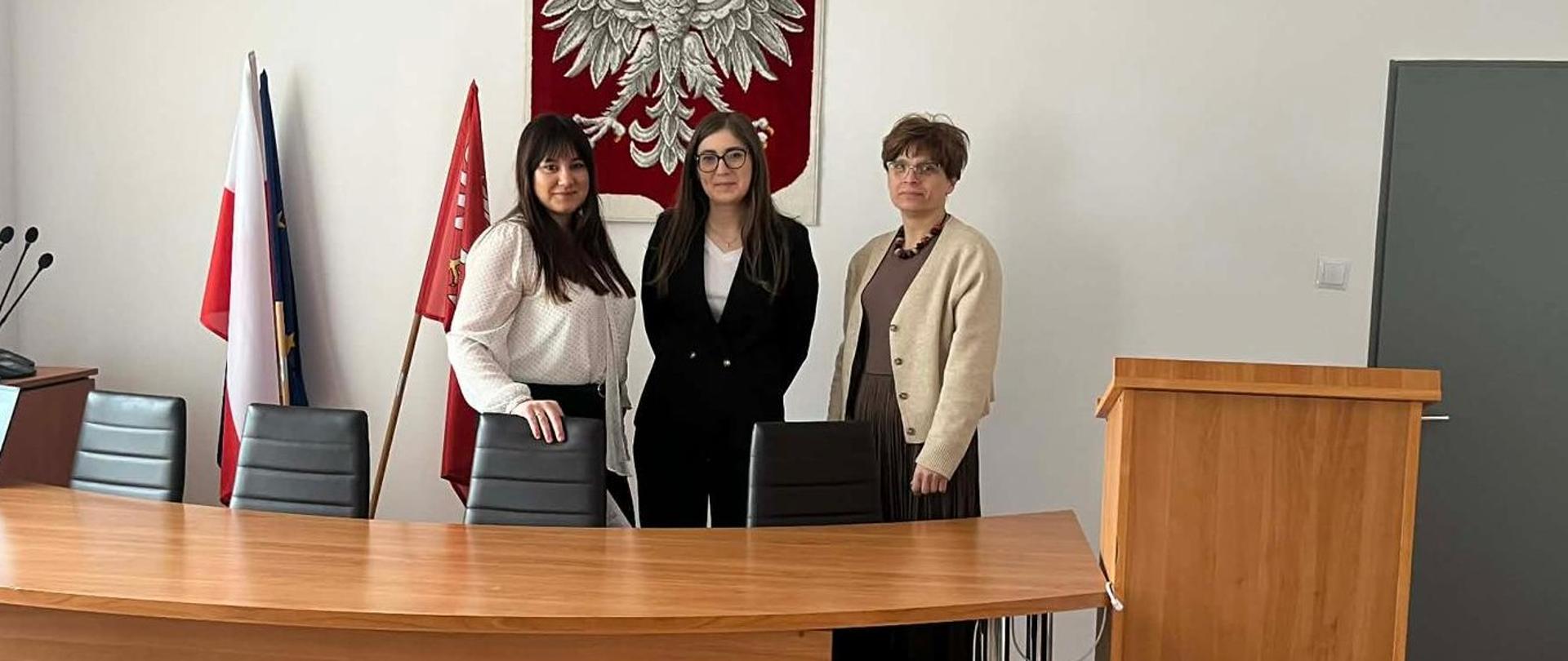 Trzy osoby stoją przy stole konferencyjnym w sali urzędowej, w tle widoczne godło Polski i flagi.