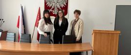 Trzy osoby stoją przy stole konferencyjnym w sali urzędowej, w tle widoczny godło Polski i flagi.