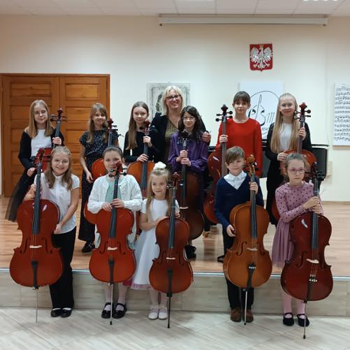 Zdjęcie przedstawia uczniów klasy wiolonczeli Pani Urszuli Właźniak stojących z wiolonczelami na scenie i przed sceną. Po lewej stronie widać grafikę czarno białą z partyturą. Za grupą wiolonczel widać grafikę z dawnym logo szkoły. Nad grafikami wisi Godło Polski.