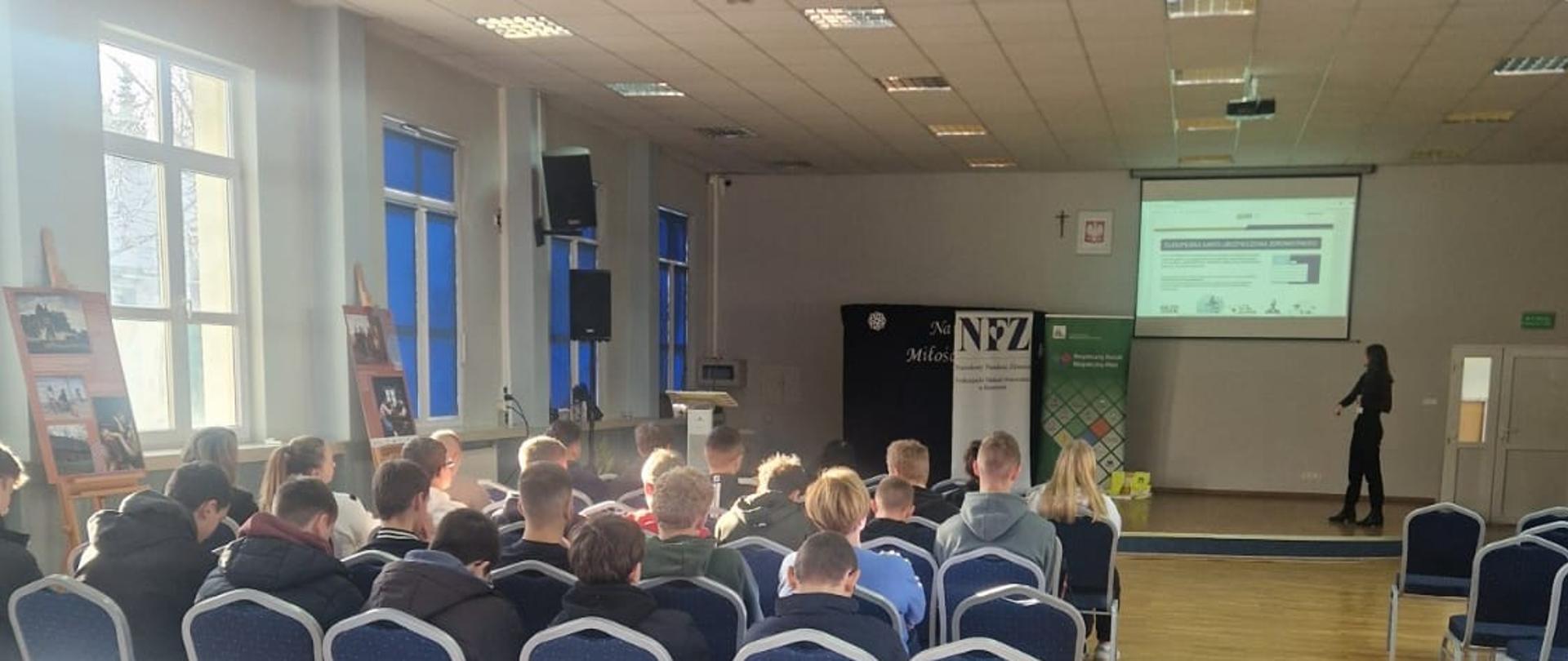 Na zdjęciu widoczna scena, na niej kobieta prowadząca prezentację. Widoczni są również siedzący uczniowie słuchający wykładu. W tle rollup NFZ oraz KRUS.
