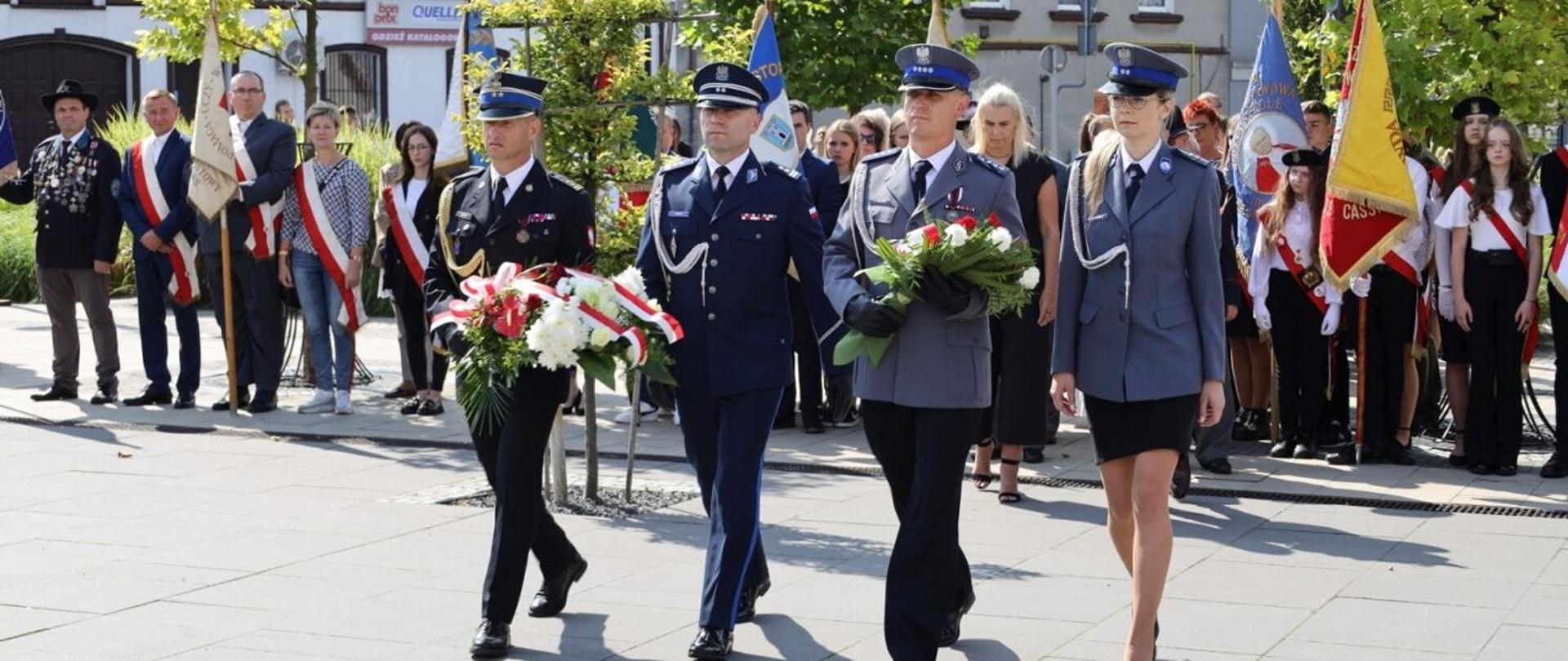 Trzech mężczyzn i jedna kobiet maszerują by złożyć wiązanki, jeden z mężczyzn ubrany w mundur wyjściowy straży pożarnej niesie wiązankę pozostałe osoby ubrane są w mundury wyjściowe policji. W tle widoczne są stojące w szeregu poczty sztandarowe