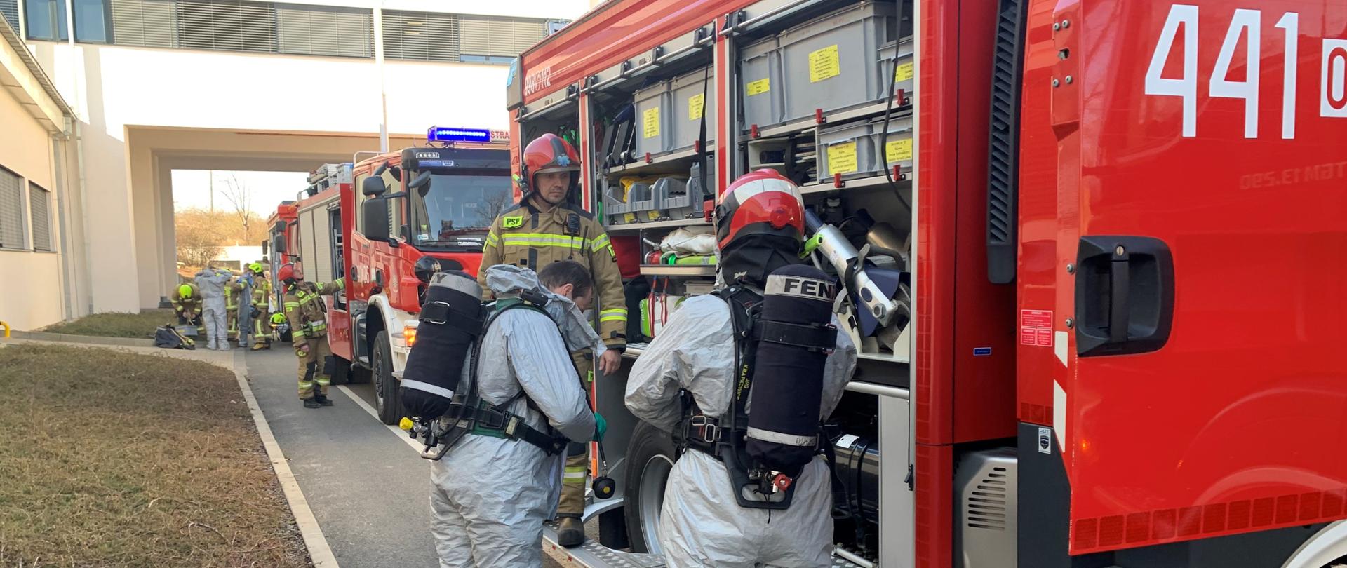 Od prawej strony widać ustawione jeden za drugim trzy samochody strażackie. Na ich tle, przy pierwszym i trzecim aucie widać przygotowujących się do akcji strażaków. Są oni w tracie zakładania ubrań ochronnych – przeciwchemicznych w kolorze szarym. Na plecach założone aparaty ochrony dróg oddechowych. Przy pierwszym aucie na pierwszym planie strażacy PSP. Dalej przy trzecim strażacy OSP. 