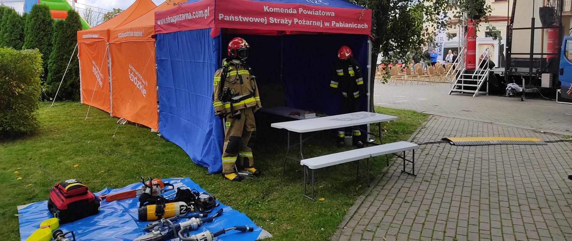 Po lewej stronie mata na której ułożony jest sprzęt ratowniczy (odcinek wężowy z prądownicą, hooligan, narzędzia hydrauliczne, aparat powietrzny oraz pilarka do drewna). Po prawej widać niebieski namiot Komendy Powiatowej PSP w Pabianicach po bokach którego stoją manekiny ubrane w strażackie ubrania specjalne. W tle po prawo scena główna.