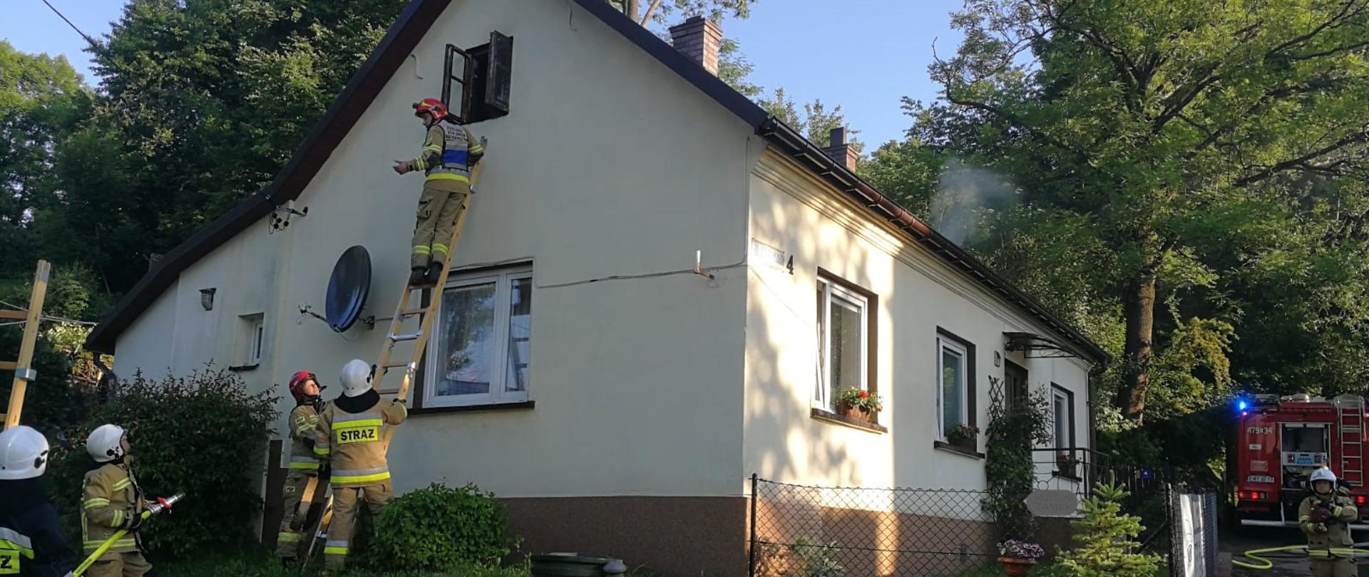 Na zdjęciu widzimy parterowy budynek mieszkalny w otoczeniu drzew i krzewów. Z lewej strony przystawiona do budynku jest drabina na której znajduje się strażak, a poniżej dwóch strażaków zabezpiecza drabinę. Obok kolejni strażacy z jednostki OSP stoją z przygotowaną i nawodnioną linią gaśniczą. Z prawej strony widoczny jest tył samochodu strażackiego.