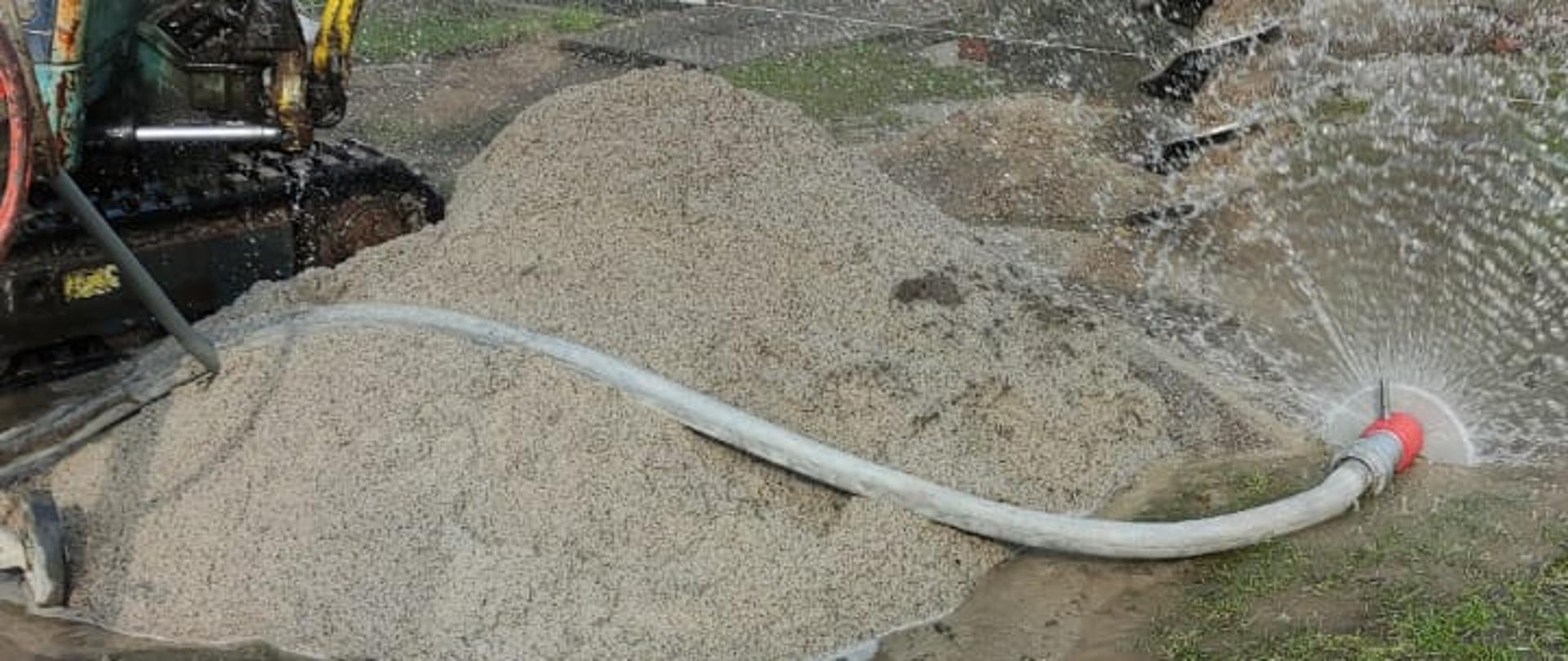 Zdjęcie przedstawiające miejsce prac ziemnych, maszynę budowlaną oraz ustawione przez strażaków urządzenie do stawiania zasłony wodnej