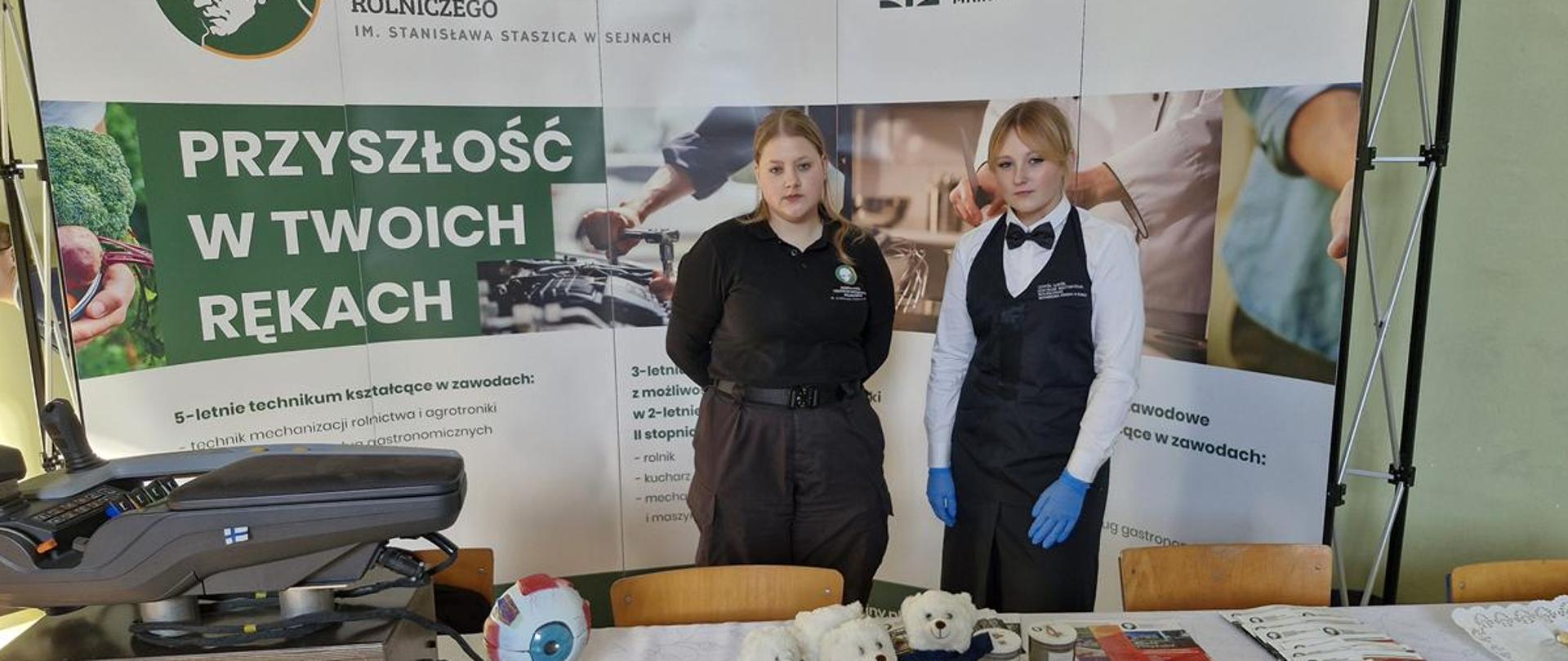 Dwie uczennice stoją na tle banera reklamującego szkołę, na stole leżą ulotki i gadżety.