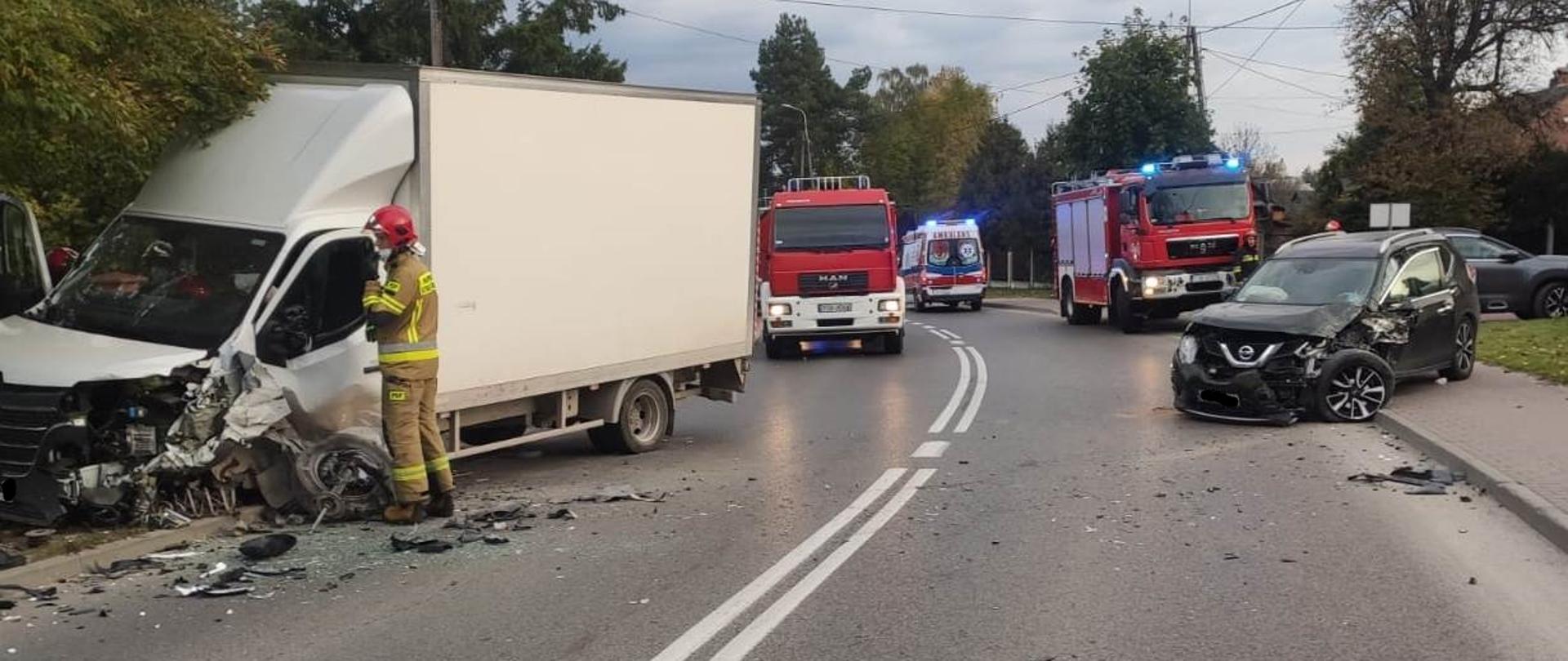 Zdjęcie obrazuje wypadek samochodowy w Suchedniowie na ul. Berezów. Samochód dostawczy zderzył się samochodem osobowym typu SUV. Zdarzenie ma miejsce na zakręcie. Bus ma uszkodzony lewy przód pojazdu, urwane koło. Osobówka ma uszkodzony przód i lewy bok pojazdu oraz lewe koło. Na jezdni leżę porozrzucane elementy uszkodzonych samochodów. Przy samochodzie dostawczym stoi strażak w umundurowaniu bojowym. Za pojazdami stoją samochody straży pożarnej z włączonymi światłami błyskowymi oraz karetka pogotowia. W tle widać drzewa.