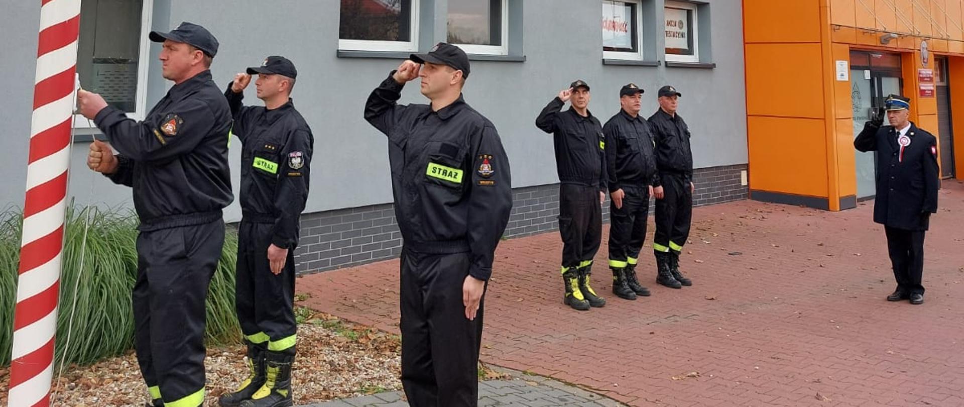 Na pierwszym planie poczet flagowy. Dwóch funkcjonariuszy oddaje honor podczas gdy jeden z strażaków wciąga flagę na maszt. Na drugim planie pozostała zmiana służbowa stojąca na baczność. Po prawej stronie Komendant Powiatowy PSP w Wolsztynie oddający honor. W Tle budynek Państwowej Straży Pożarnej w Wolsztynie. 