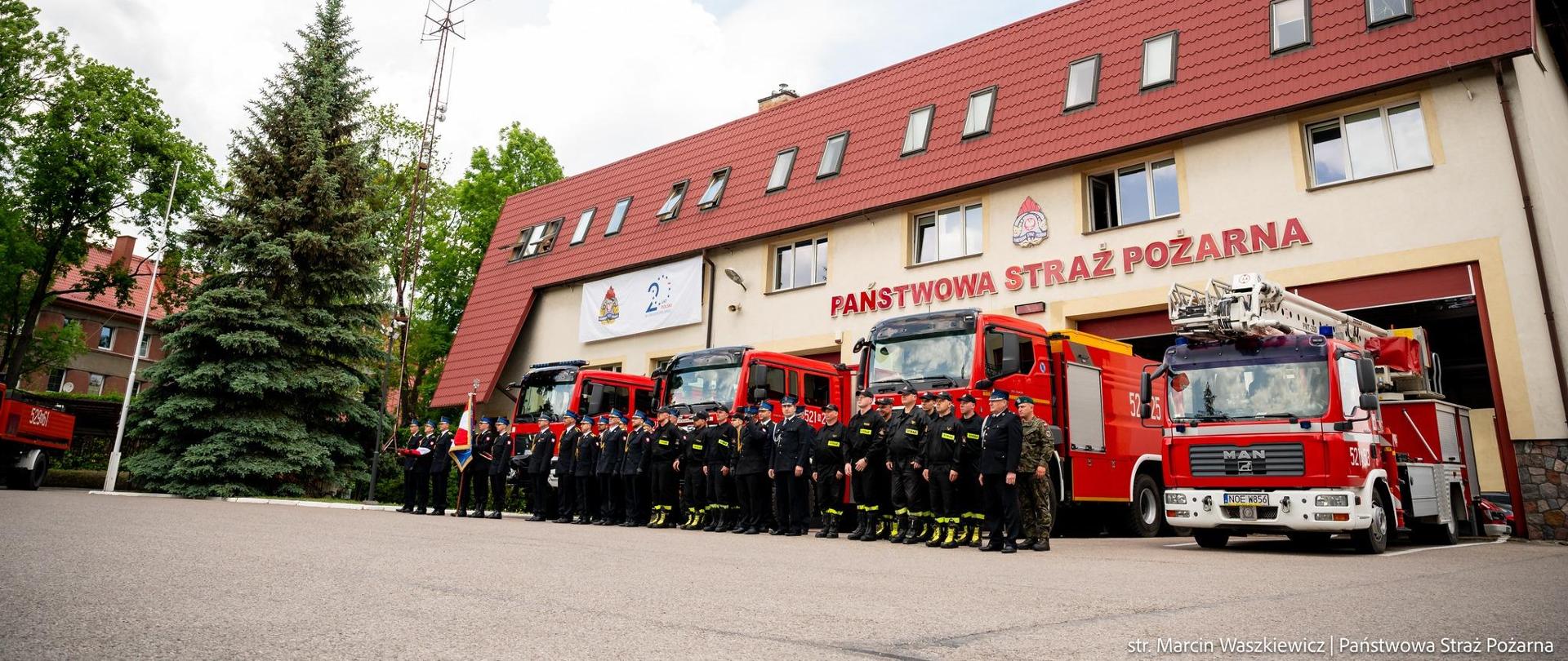 Dzień Strażaka - strażacy na tle pojazdów i budynku komendy