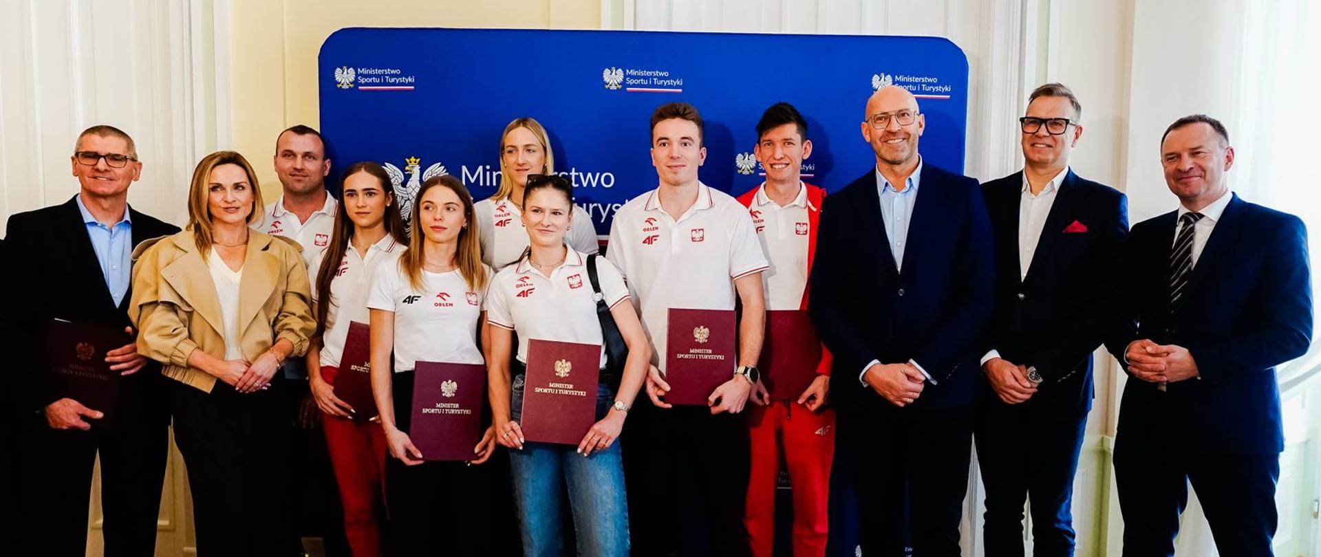 Spotkanie ministra Rutnickiego z uczestnikami HMŚ w Toruniu. Zawodnicy w białych strojach stoją na tle granatowej ścianki z logiem ministerstwa. Po prawej stronie stoją ministrowie w garniturach