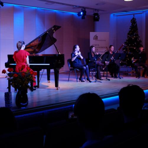 zdjęcie przedstawia kwintet dęty i solistkę przy fortepianie podczas wykonania utworu na scenie sali koncertowej, w tle baner szkoły