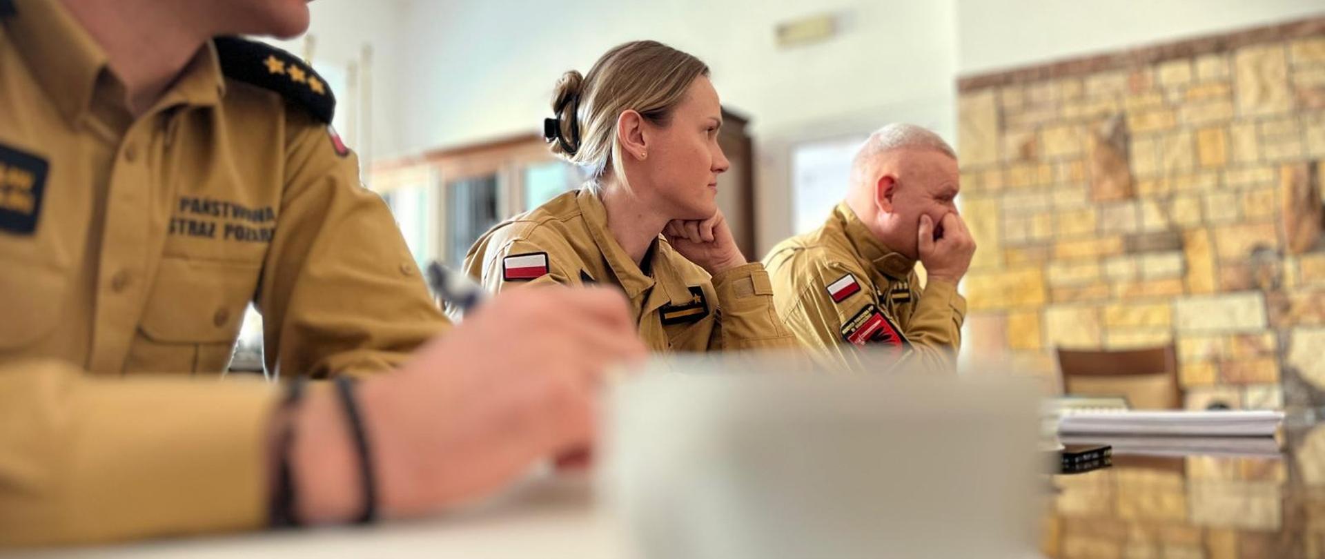 Przy stole trzech strażaków, w środku strażak - kobieta. W mundurze służbowym, piaskowym. W tle fragment ściany z godłem narodowym. Z przodu rozmazany widok filiżanki.