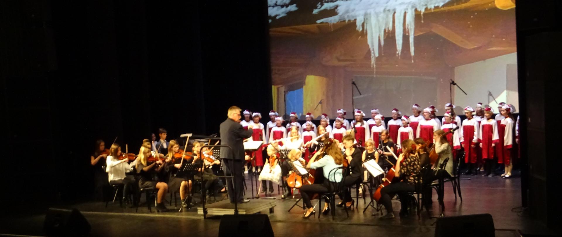 Zdjęcie przedstawia występ chóru w strojach biało-czerwonych i orkiestry na tle scenerii zimowej