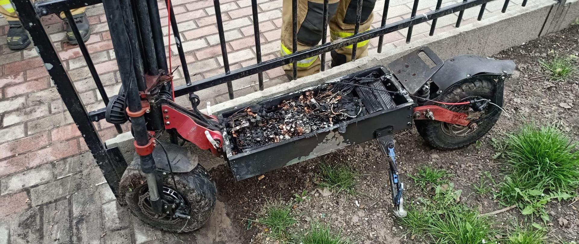 Na trawniku, przy płocie stoi spalona hulajnoga elektryczna. W tle widoczna sylwetka strażaka.