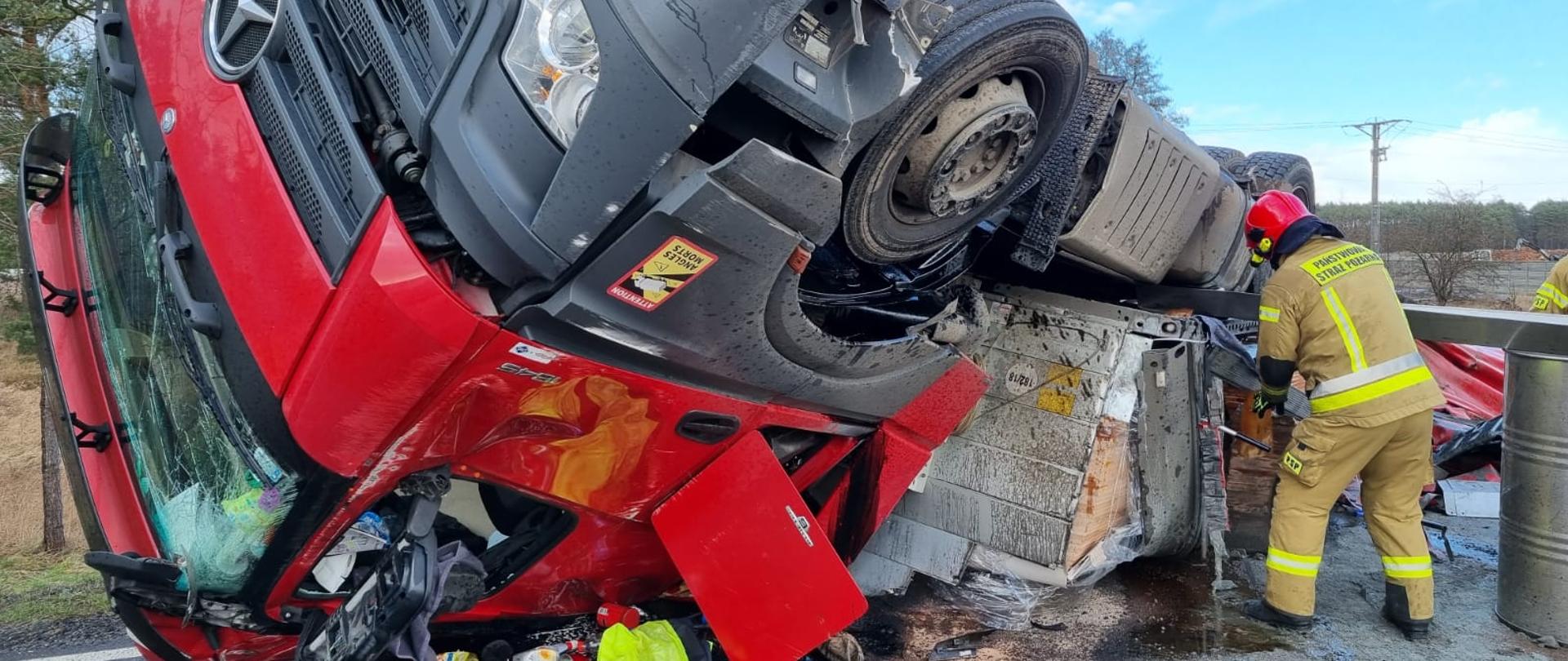 Samochód ciężarowy przewrócony na dachu leży na drodze obok strażak prowadzi działania.