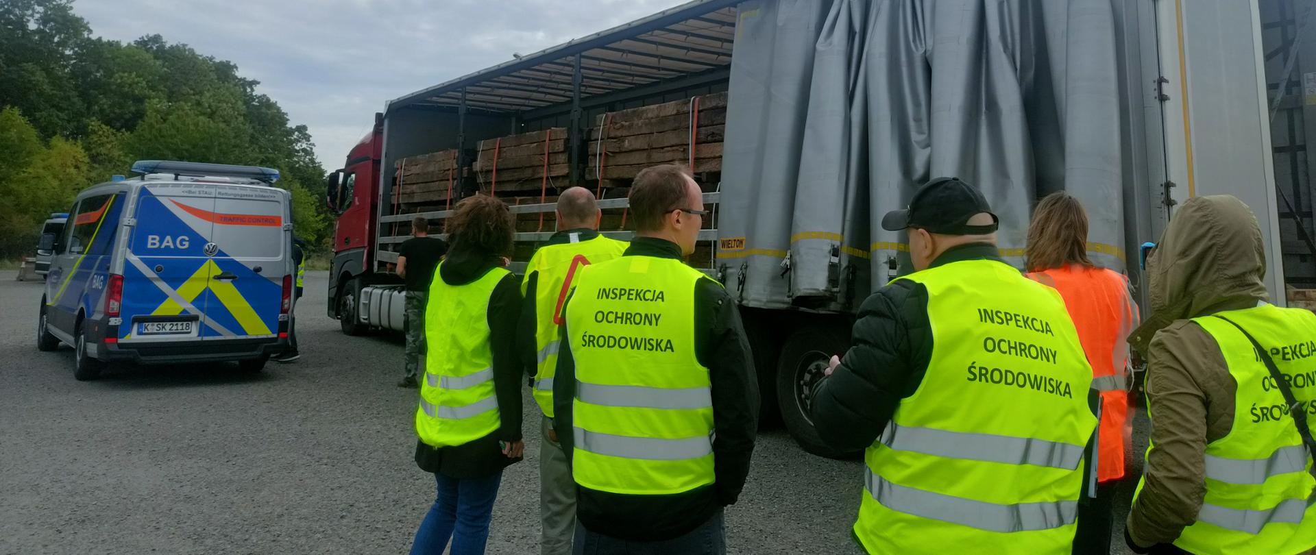 Meżczyźni i kobiety w odblaskowych kamizelkach stoją przez naczepą tira, która jest odsłonięta. W środku naczepy znajdują się betonowe słupki. Obok tira stoi szaro-niebieski samochód niemieckich służb.