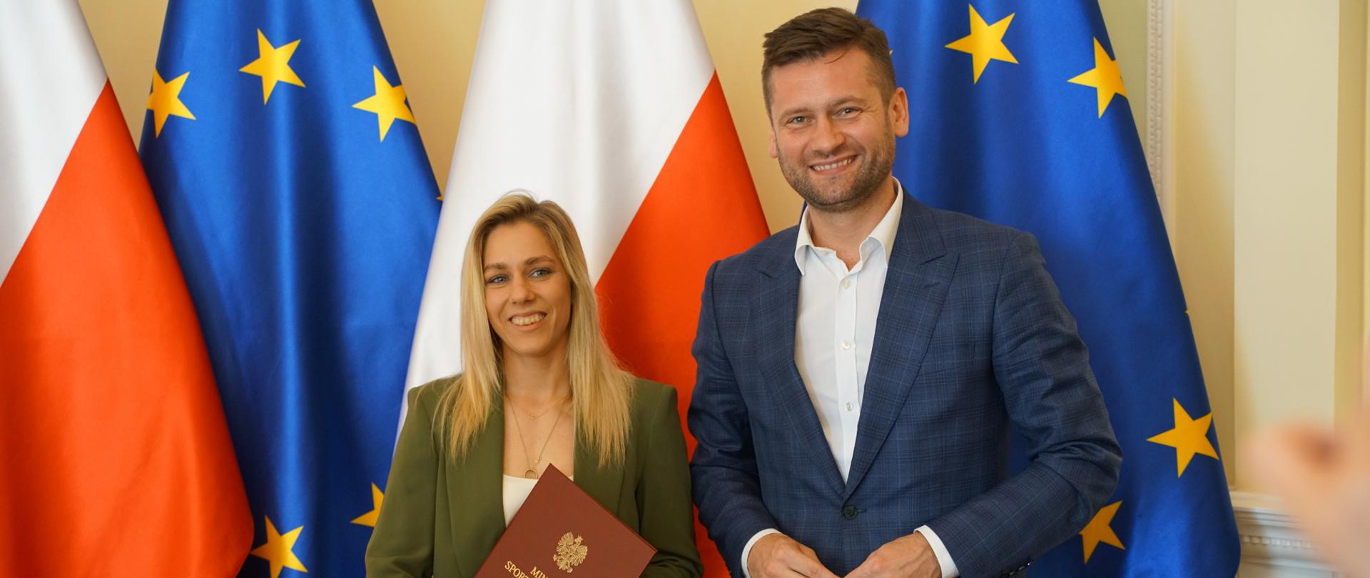 Minister Kamil Bortniczuk pozuje na tle flagi Polski i Unii Europejski z Aleksandrą Mirosław, reprezentantką Polski we wspinaczce sportowej. Kobieta trzyma torebkę z napisem Ministerstwo Sportu i Turystyki oraz bordową teczkę.