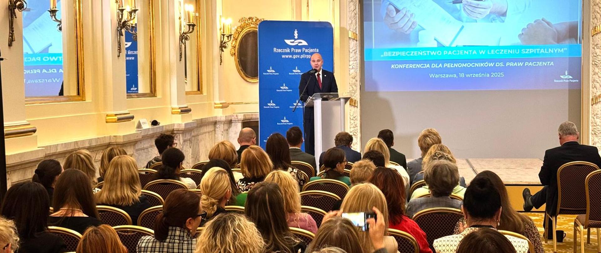 Zdjęcie przedstawia elegancką salę, przypominającą pałacowe wnętrza. na pierwszym planie krzesła i osoby na nich siedzące, widoczne tyłem. Na scenie za mównicą przemawia Rzecznik Praw Pacjenta Bartłomiej Chmielowiec, jedyna osoba widoczna przodem. W tle na dużym ekranie pierwszy slajd prezentacji Bezpieczeństwo Pacjenta w Leczeniu szpitalnym.