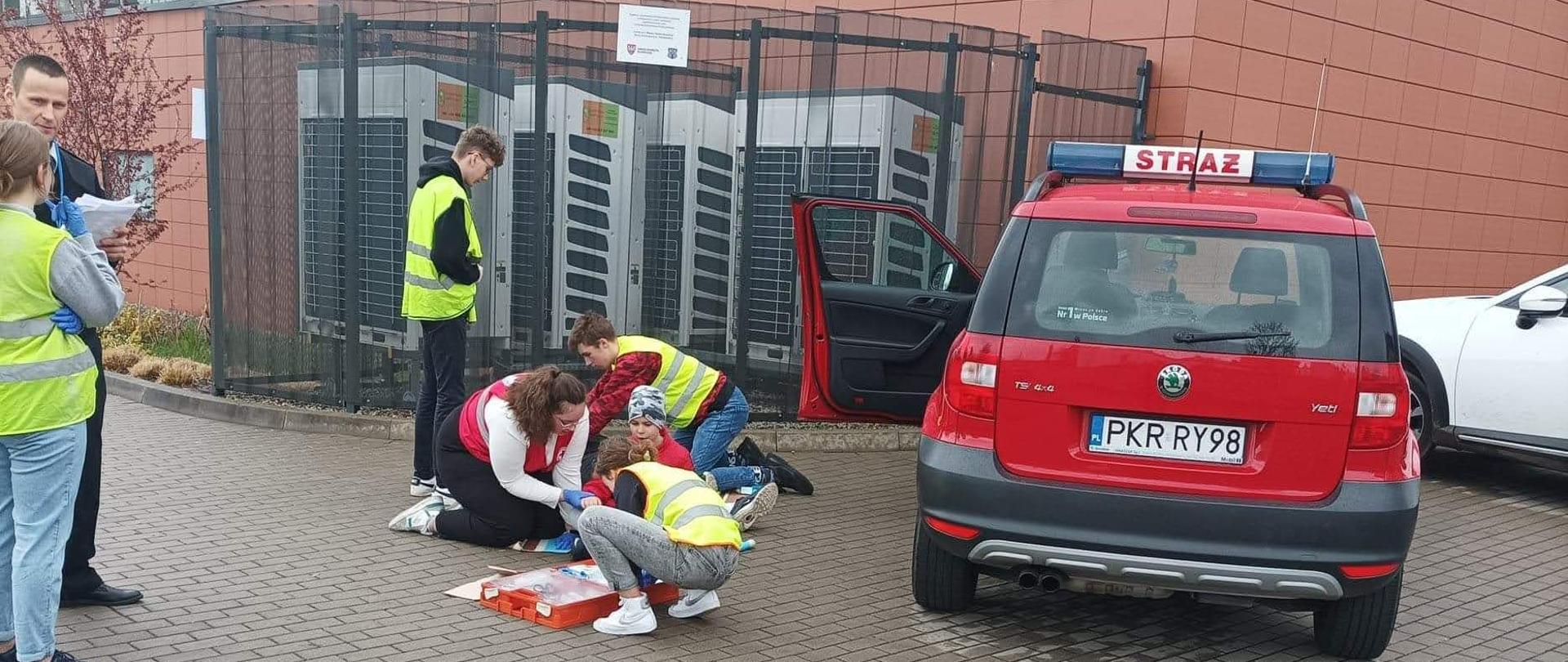 Pozoracja wypadku samochodowego. Przed pojazdem stoi trójkąt ostrzegawczy. Zespół udziela pomocy medycznej obok pojazdu. Całe zagadnienie ocenia strażak .