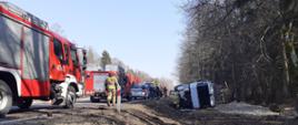 Po lewej na jezdni stoją samochody służb ratowniczych. Po prawej w tle leży przewrócony na bok samochód ciężarowy w przydrożnym rowie. Po prawej las.