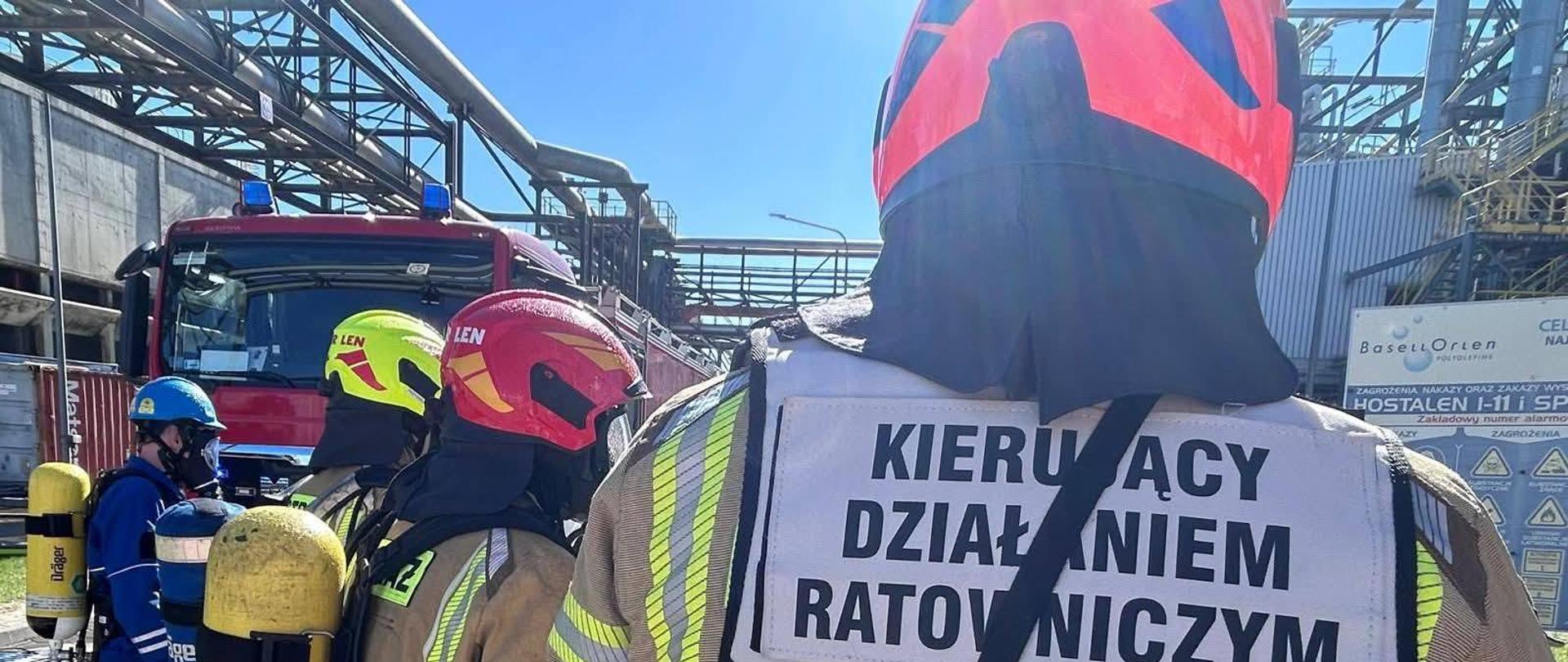 Na zdjęciu widać grupę strażaków w ubraniach ochronnych, wyposażonych w aparaty powietrzne z żółtymi butlami. Jeden z nich, na pierwszym planie, ma na plecach kamizelkę odblaskową z napisem „KIERUJĄCY DZIAŁANIEM RATOWNICZYM”, wskazującą, że pełni funkcję dowódcy akcji. Obok stoją strażacy w czerwonym i żółtym hełmie z napisem „ORLEN”, a w tle widoczny jest pracownik w niebieskim kombinezonie i hełmie ochronnym. W tle stoi czerwony wóz strażacki z włączonymi światłami oraz zabudowania przemysłowe z instalacjami rurowymi i tablicami ostrzegawczymi. Akcja odbywa się przy dobrej pogodzie, w pełnym słońcu.